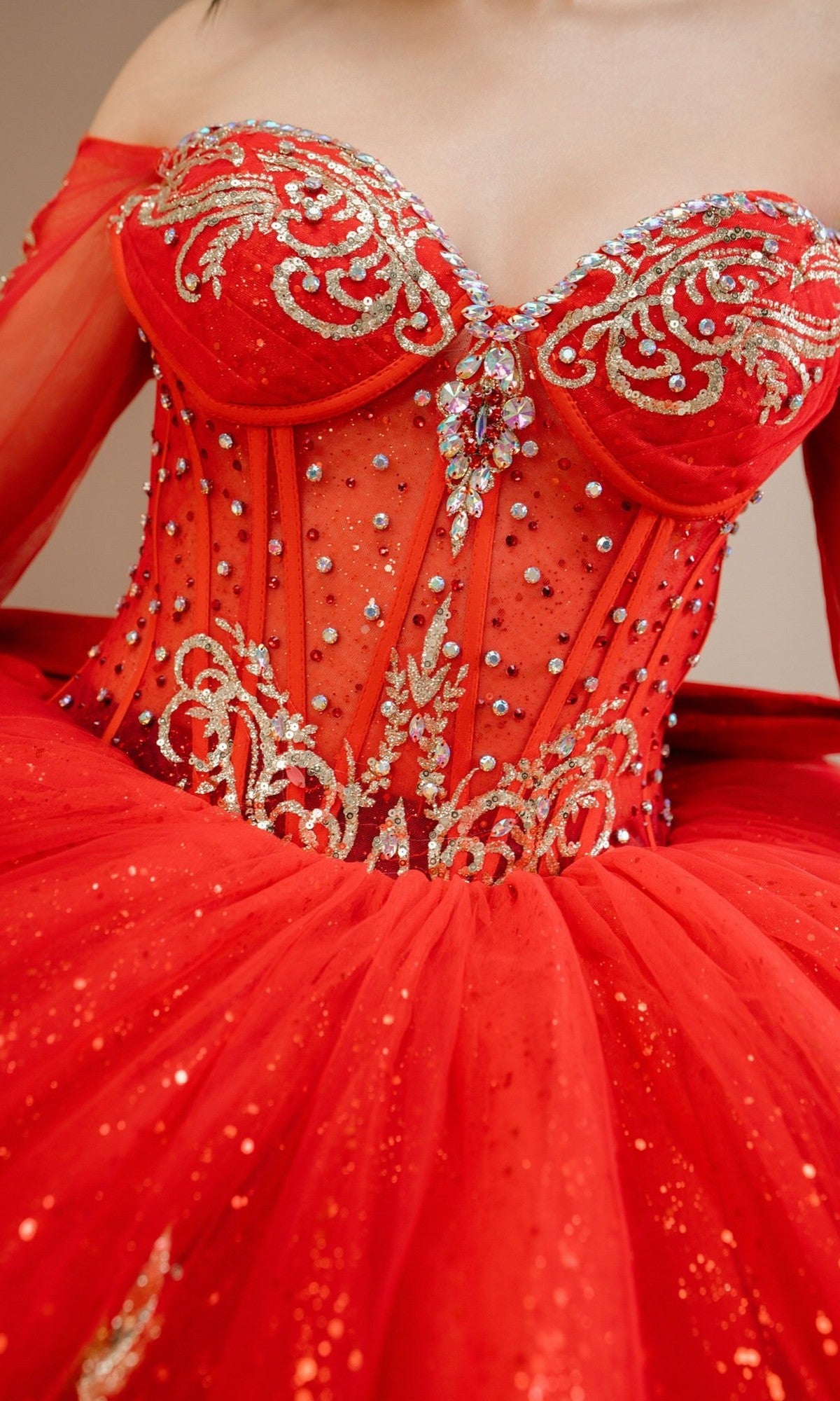 A close-up of a person wearing the Quinceanera Dress 8545JQ By Cinderella Couture, a red ball gown with an off-the-shoulder sweetheart neckline, gold embroidery, and sparkling beaded sequin embellishments.